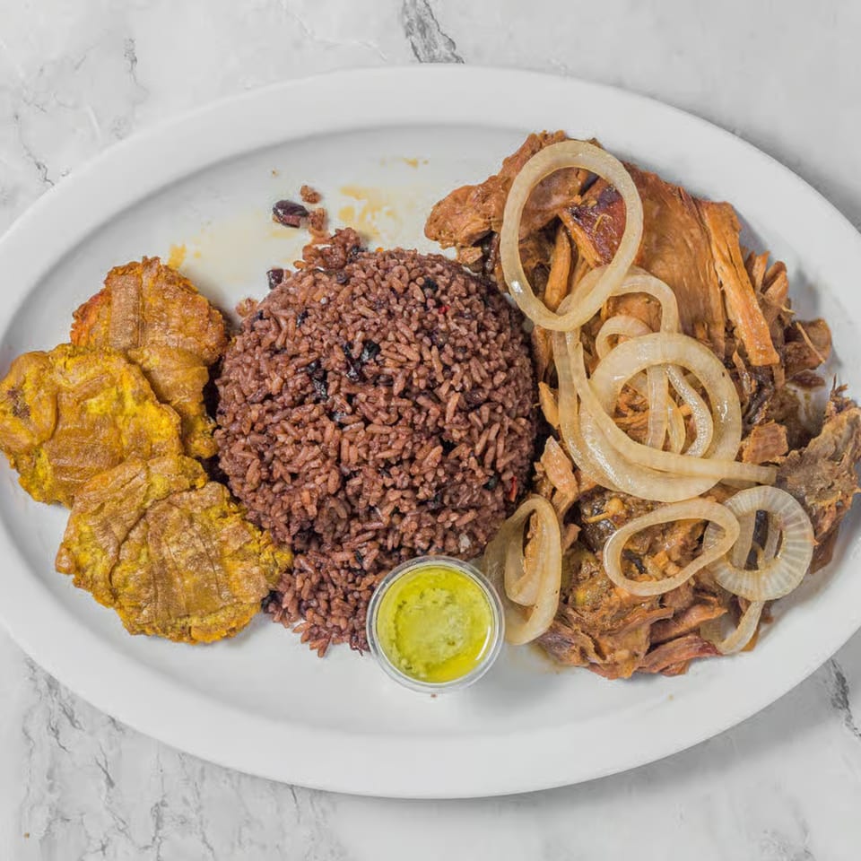 A white platter holds a meal of brown rice, fried plantains, shredded meat with onions, and a small cup of green sauce.