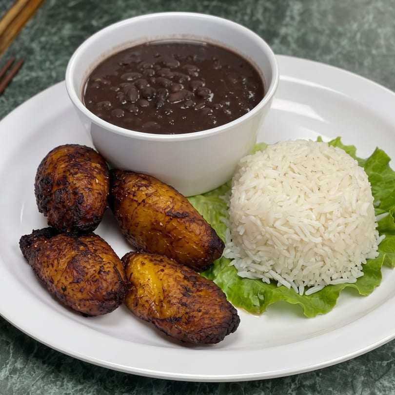 A white plate with a bowl of black beans, white rice on lettuce, and four fried plantains.