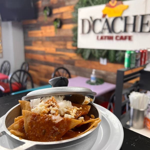 Chilaquiles with red sauce and cheese in a metal pot, with D'Cache Latin Cafe sign in background.