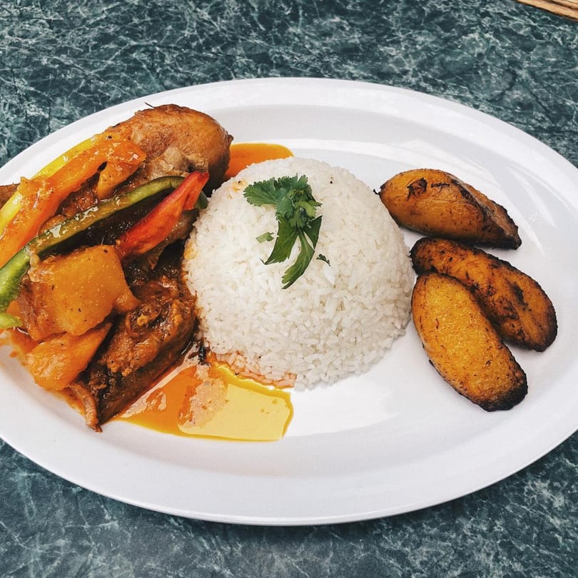 A white plate with chicken stew, white rice, and three fried plantains on a dark marbled table.
