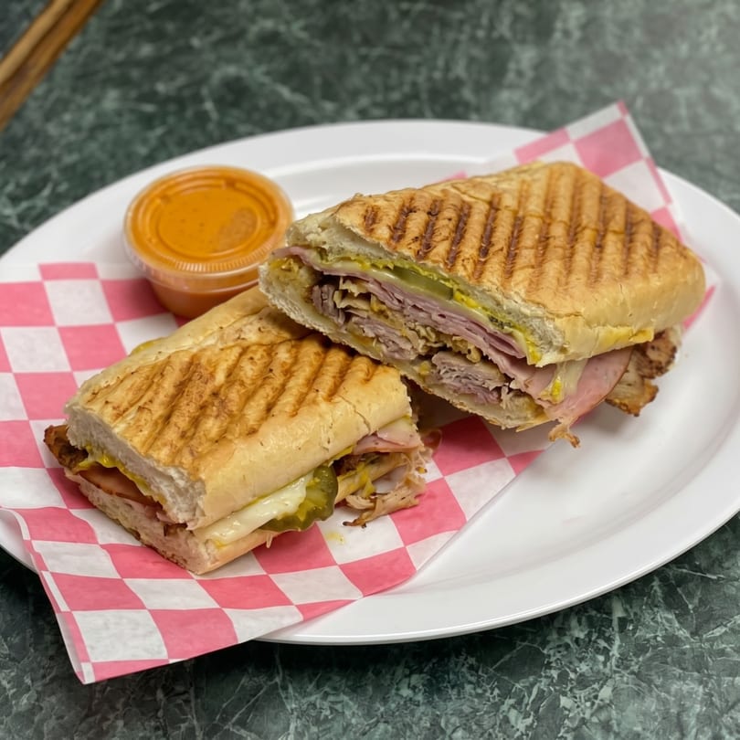 Grilled Cuban sandwich, cut in half, with a side of orange sauce on a white plate.
